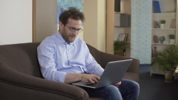 Hombre con anteojos sentado en el sofá y compras en línea en el ordenador portátil, tecnologías — Vídeos de Stock