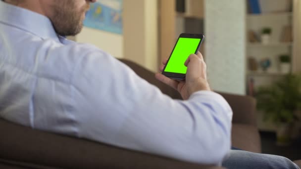 Bearded male sitting on sofa and scrolling on cellphone with green screen, app — Stock Video