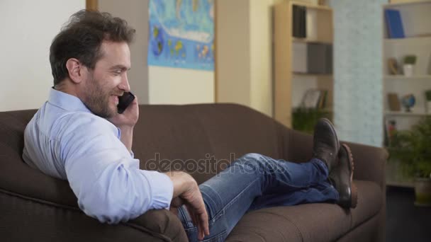 Hombre feliz charlando con el mejor amigo en el teléfono inteligente, sentado en el sofá en casa — Vídeos de Stock