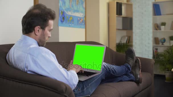 Hombre sentado en el sofá y viendo la película favorita en el ordenador portátil con pantalla verde — Vídeo de stock
