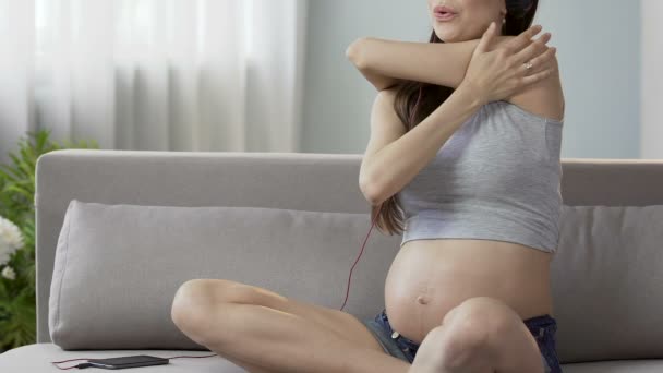 Μελλοντική μητέρα κάθεται στον καναπέ στα ακουστικά, τεντώνοντας το ανώτερο σώμα, περιποίηση σώματος — Αρχείο Βίντεο