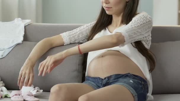 Zwangere vrouw zittend op de Bank nemen baby schoenen om te kijken naar hen, baby mode — Stockvideo