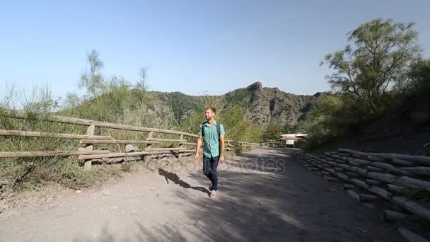 Unga manliga backpacker kommer uppförsbacke längs marken road i bergen på solig dag — Stockvideo