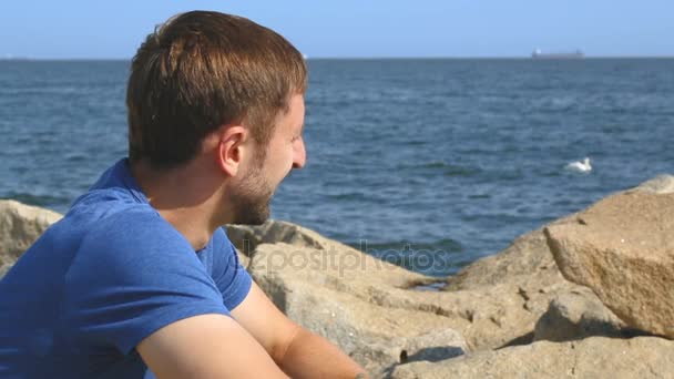 Feliz joven macho sentado en piedras en el terraplén y disfrutando de hermosos paisajes marinos — Vídeos de Stock