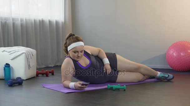 Lusta kövér lány feküdt szőnyeg, görgetés a szociális média modul helyett edzés — Stock videók