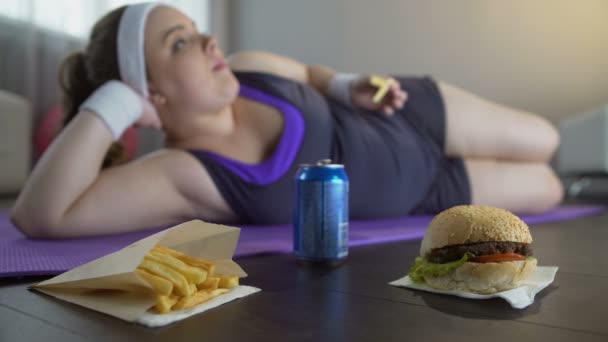 Dama perezosa en ropa deportiva acostada en la esterilla de yoga comiendo comida chatarra en lugar de entrenamiento — Vídeos de Stock