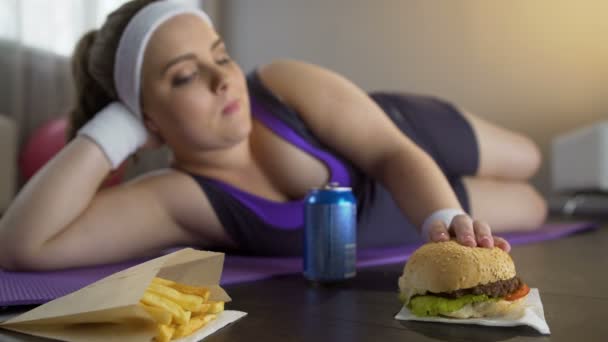 Счастливая самоуверенная тучная девушка ест жирный бургер вместо спортивных тренировок — стоковое видео