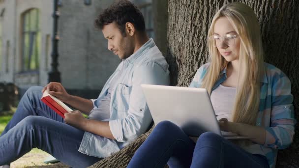 Ragazza intelligente in occhiali che lavora su computer portatile, sorridendo a ragazzo romantico con libro — Video Stock