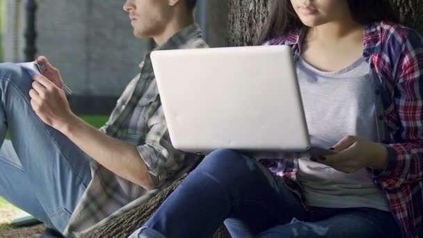 Couple of students doing their homework, girl working on laptop under tree — Stock Video