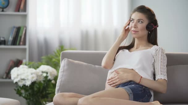 Молодая беременная женщина в наушниках слушает музыку, нежно трогает живот — стоковое видео