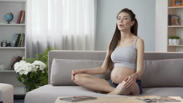 Будущая мать сидит на диване, делает йогу для беременных дыхательные упражнения, расслабляет — стоковое видео