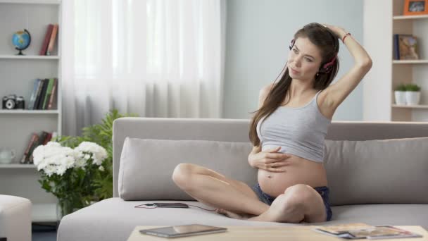 Jeune femme enceinte assise sur le canapé dans un casque, faisant des exercices d'étirement — Video