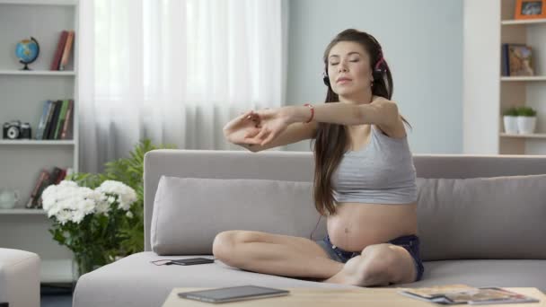 Будущая мама сидит на диване в наушниках, растягивая спину, аудио тренировки — стоковое видео