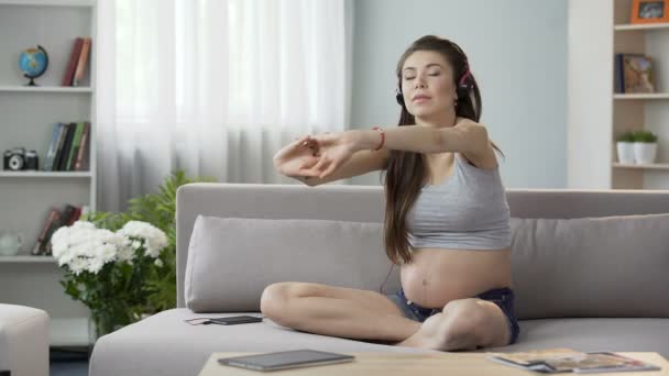 Futura madre sentada en el sofá en los auriculares, estiramiento hacia atrás, entrenamiento de audio — Vídeo de stock