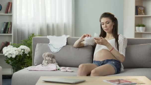 Jovem mulher grávida dando uma olhada em crianças roupas no sofá, moda bebê — Vídeo de Stock