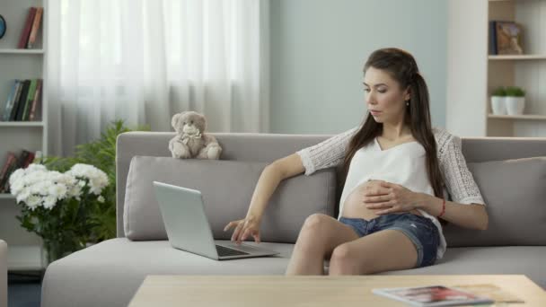 Очікування мати сидить на дивані і використовує ноутбук, інформацію про вагітність — стокове відео