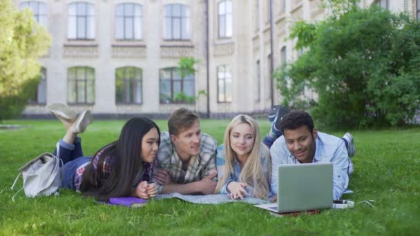 Multi-ethnic students lying on grass and watching funny video on laptop, friends — Stock Video