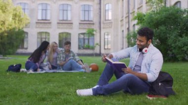 Çimenlerin üzerinde oturan ve ilginç bir kitap okuma mutlu biracial erkek öğrenci
