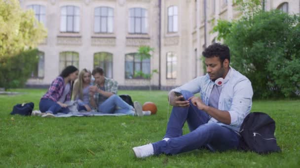 Junge hispanische männliche Student sitzt auf Gras und blättert auf Handy, entspannen — Stockvideo