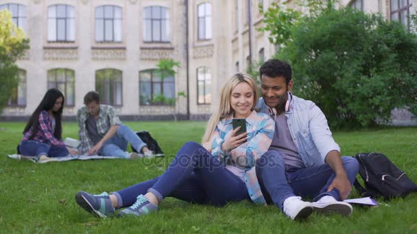 Amar a un par de estudiantes sentados en el césped y viendo video en el teléfono inteligente — Vídeos de Stock