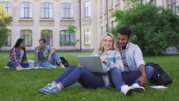 Estudiantes multiétnicos sentados en la hierba y viendo videos divertidos en el portátil — Vídeos de Stock