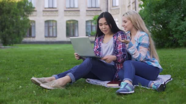 Две студентки сидят на траве и выбирают новые платья на ноутбуке, устройстве — стоковое видео
