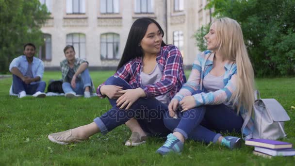 Mujeres sentadas en la hierba, hablando y sonriendo, chicos mirando a las chicas, coqueteando — Vídeo de stock