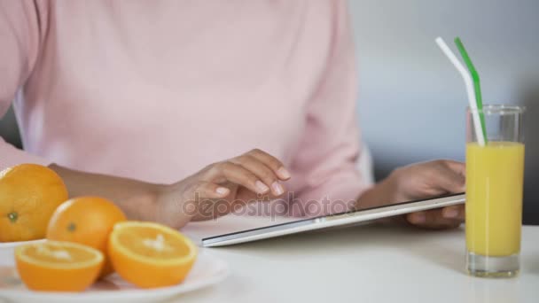 Змішана расова леді, використовуючи планшет, апельсини та сік на столі, пошук статті про здоров'я — стокове відео