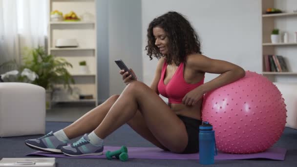 Biracial vrouw in sportschool outfit zittend op de vloer, met behulp van de mobiele telefoon, toepassing — Stockvideo