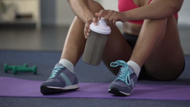 Mujer joven de raza mixta sentada en el suelo en ropa deportiva, bebiendo proteínas — Vídeos de Stock