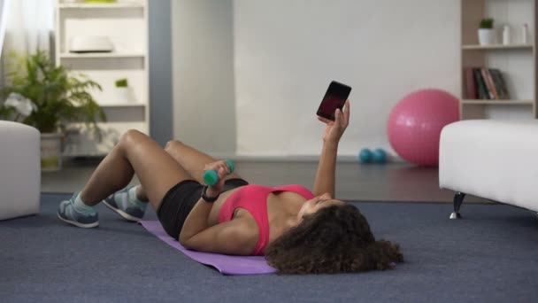 Femme couchée sur le sol, haltère de levage avec une main et en utilisant un téléphone mobile — Video