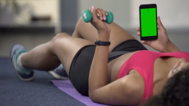 Perempuan muda ras campuran menggunakan telepon dengan layar hijau, lengan lentur dengan dumbbell — Stok Video