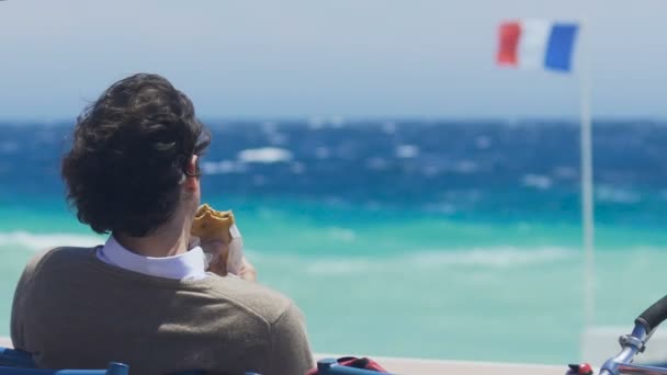 Unavený turista sedí na pláži pozoroval vlny a jíst sendvič, pomalý pohyb — Stock video