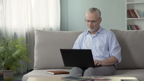 Saudável macho sênior sentado no sofá e visualização de fotos no namoro site, laptop — Vídeo de Stock