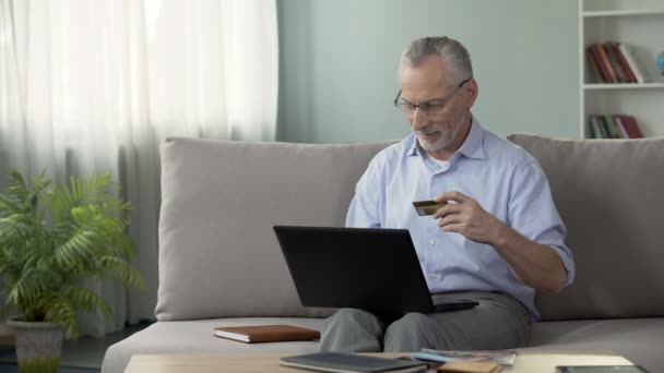 Пенсионер сидит на диване и вставляет номер карты на ноутбук, онлайн покупки — стоковое видео