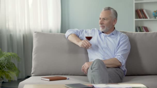 Varón pensativo en sus cincuenta años sentado en el sofá y disfrutando del sabor del vino tinto — Vídeo de stock