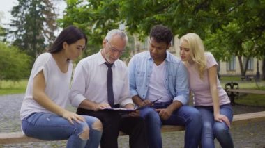 Mutlu öğretmen ve çok etnik gruptan oluşan öğrenciler banktaoturuyor, kameraya gülümseyen