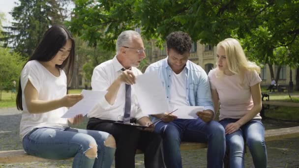 Professor qualificado dando testes para estudantes multirraciais no parque perto da universidade — Vídeo de Stock