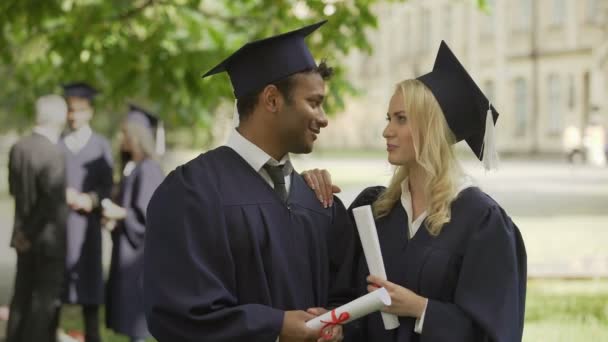 Graduados en regalia académica hablando, chica arreglando corbata, chico besándola en la mejilla — Vídeo de stock