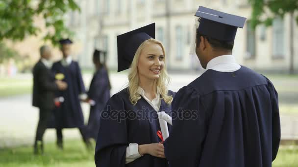 Graduada en sombrero cuadrado con diploma hablando con compañera de clase, día de la convocatoria — Vídeo de stock