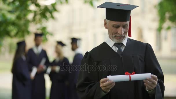 Senior man in academic regalia holding diploma, education at any age, new degree — Stock Video