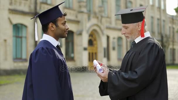 Выдающийся профессор вручает диплом студенту мужского пола, пожимающему руку в день созыва — стоковое видео