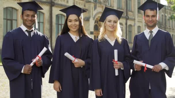 Graduados universitarios en regalia académica que poseen diplomas, poniendo pulgares hacia arriba — Vídeo de stock