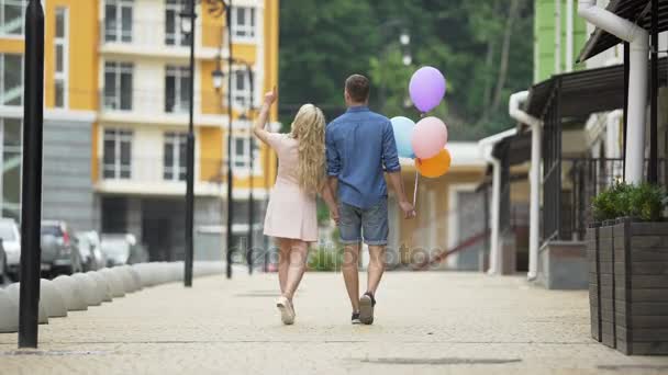 Молодая пара, идущая по улице, держащая за руки парня, держащего воздушные шары, романтичная — стоковое видео