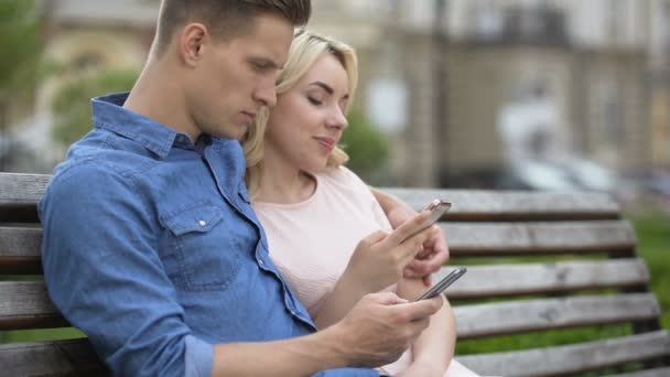 Młoda para siedzi na ławce i za pomocą telefonów komórkowych, sieci społecznych, rozproszenia — Wideo stockowe