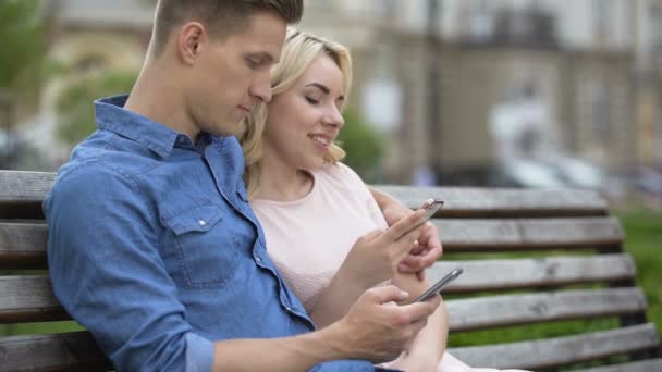 Masculino e feminino sentado no banco com telefones celulares, cara beijando menina na bochecha — Vídeo de Stock