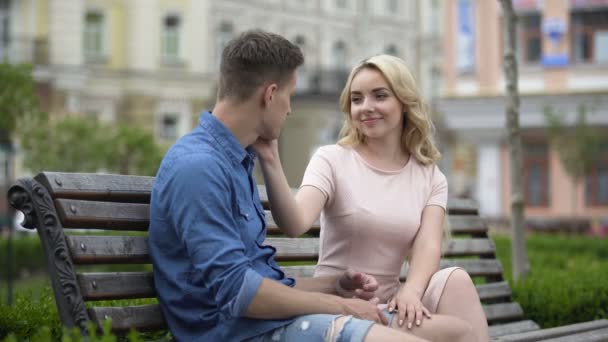 Молодая пара сидит на скамейке, парень смотрит на девушку и ласкает ее, нежность — стоковое видео