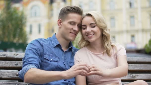 Junges Paar, das eng beieinander sitzt, ein fingerförmiges Herz hochhält und sich küsst — Stockvideo