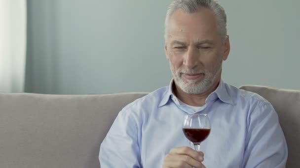 Hombre de pelo gris sentado en el sofá y sosteniendo una copa de vino, disfrutando de su olor — Vídeo de stock