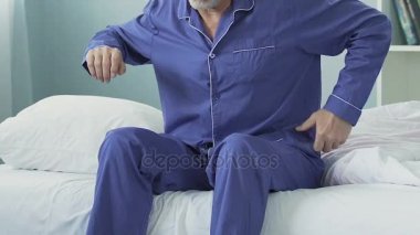 Yatağın kenarında oturan, germe ve ani bel ağrısı olan yaşlı adam